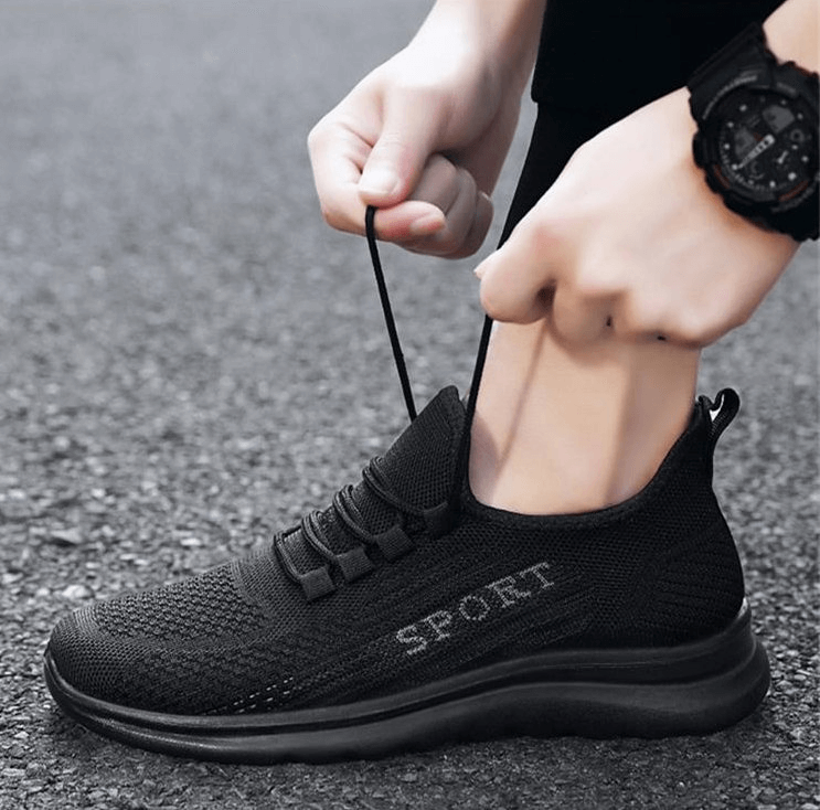Person tying black breathable lace up sneakers for men, featuring mesh upper and stylish design on pavement.